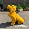 Carica l'immagine nel visualizzatore della galleria, A large yellow plush dog statue or mascot sitting on an outdoor paved surface, with a person partially visible in the upper left and a brick wall and plants in the background. A watermark reading mcsdino.