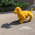 Carica l'immagine nel visualizzatore della galleria, A yellow plush dog toy standing on an outdoor paved surface with green plants and a building wall in the background. White text at the bottom right reads 'mcsdino.