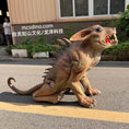 Carica l'immagine nel visualizzatore della galleria, A realistic dinosaur-like creature model or prop with a textured brown and tan skin, large ears, and an open mouth showing red interior, photographed outdoors on a paved surface in front of a building. A person is partially visible in the background, and a watermark reading 'mcsdino.