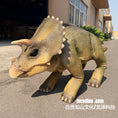 Load image into Gallery viewer, A realistic toy or model of a Triceratops dinosaur with textured tan and olive-green skin, standing on an outdoor paved surface near a building.