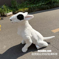 Load image into Gallery viewer, A white sheep-like plush toy or stuffed animal with a large black eye patch sits on an outdoor paved surface. The background shows plants, a brick wall, and a street setting.
