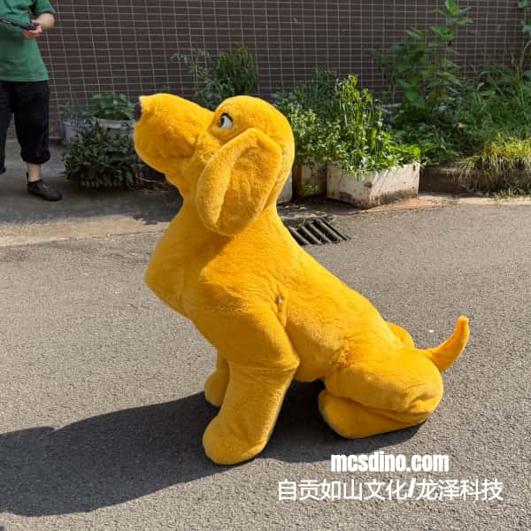 A large yellow plush dog statue or mascot sitting on an outdoor paved surface, with a person partially visible in the upper left and a brick wall and plants in the background. A watermark reading mcsdino.