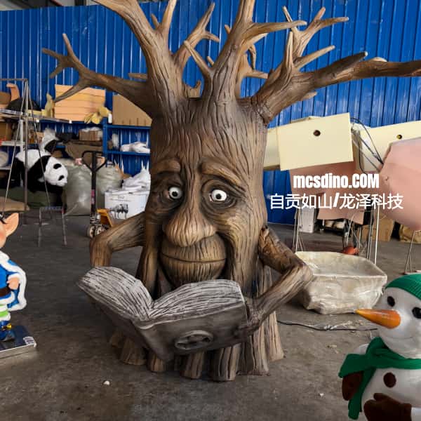 A large tree-shaped fantasy statue with a carved face reading a book, photographed in a workshop or production space with blue corrugated walls and other decorative items around it.