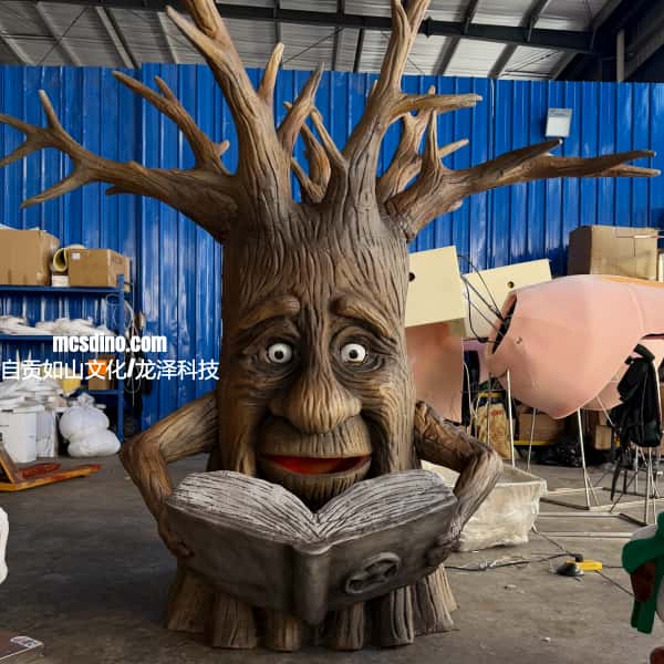 A large tree-like book character sculpture with a funny face, holding an open book, photographed in a workshop or studio setting with blue corrugated walls and construction materials in the background.
