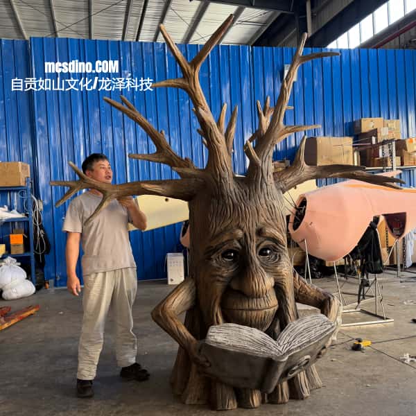 A large carved tree-like sculpture with antler-like branches and a face reading an open book, shown in a workshop next to a man. The background includes blue corrugated metal walls, boxes, and studio equipment.