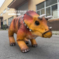 Load image into Gallery viewer, A realistic toy or model of a Triceratops dinosaur with orange, brown, and red textured skin, standing on an asphalt surface in front of a building.
