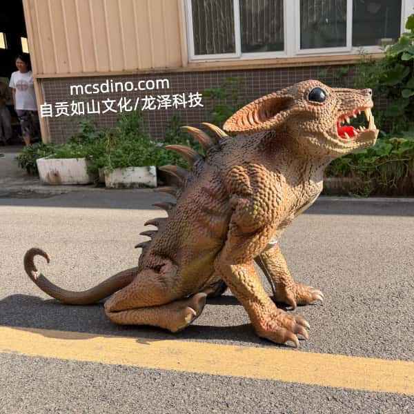 A realistic dinosaur-like creature model or prop with a textured brown and tan skin, large ears, and an open mouth showing red interior, photographed outdoors on a paved surface in front of a building. A person is partially visible in the background, and a watermark reading 'mcsdino.