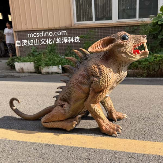 A realistic dinosaur-like creature model or prop with a textured brown and tan skin, large ears, and an open mouth showing red interior, photographed outdoors on a paved surface in front of a building. A person is partially visible in the background, and a watermark reading 'mcsdino.
