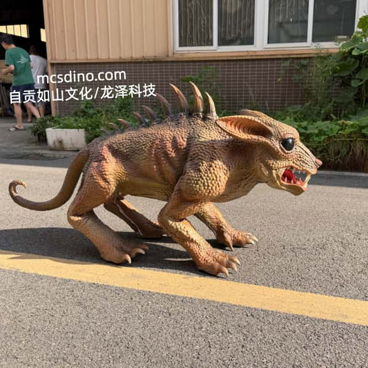 A realistic dinosaur toy or prop dinosaur standing on an outdoor paved surface in front of a building, with visible people and plants in the background. The dinosaur is tan and brown with darker ridged spikes along its back and an open mouth showing red teeth.