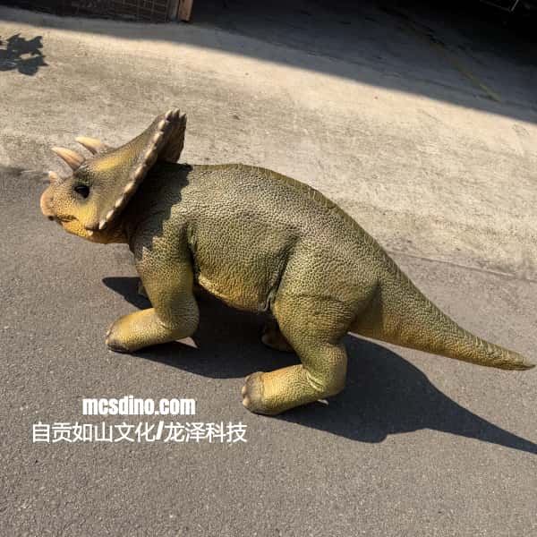 A realistic toy or model Triceratops dinosaur with a textured olive-green and tan body, standing on asphalt outdoors. The watermark 'mcsdino.