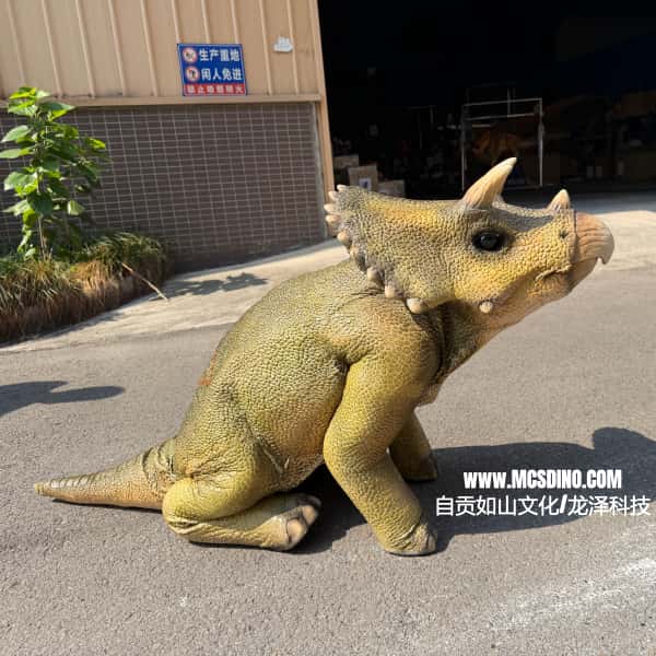 A realistic baby Triceratops dinosaur toy or prop shown outdoors on asphalt in front of a building. The toy has textured olive-green and tan skin, small horns, and a horned head crest.